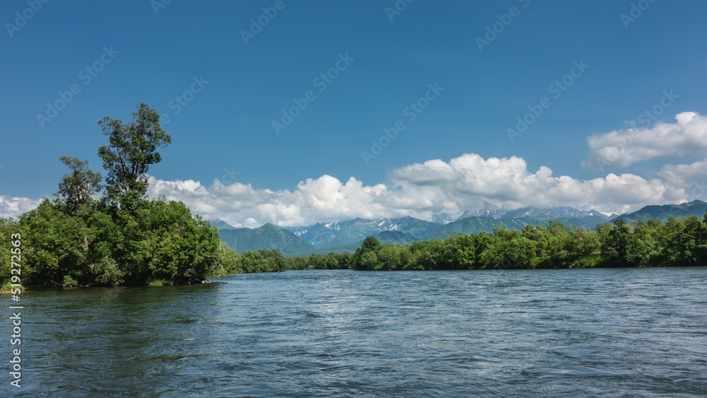 The bed of the calm blue river bends between the green banks. Mountains on the background of azure sky and clouds. Kamchatka. River Bystraya