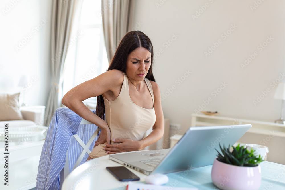 Businesswoman Holding Her Back While Working On Laptop At Home Office Desk. young woman, stretching, sitting feeling her back tired after working at laptop, small home office interior. Businesswoman Holding Her Back While Working On Laptop At Home Office Desk. young woman, stretching, sitting feeling her back tired after working at laptop, small home office interior.