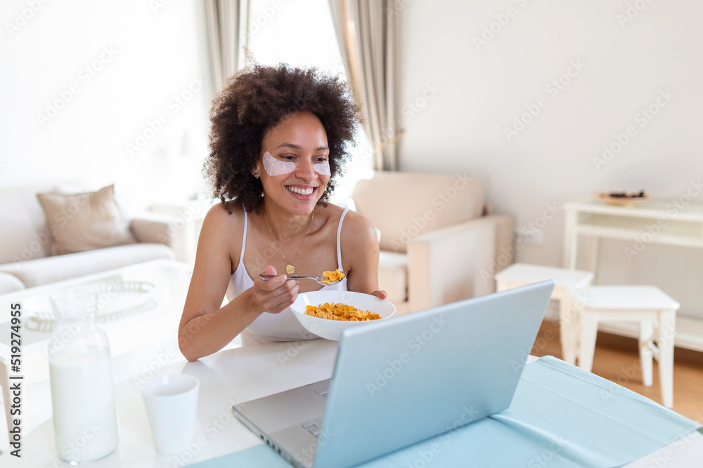 young amazing woman sitting indoors at the table with laptop holding corn flakes. Looking at laptop computer and talking to her friends via video call.