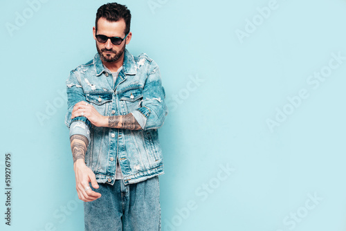 Portrait of handsome confident stylish hipster lambersexual model. Sexy man dressed in jeans jacket. Fashion male isolated on blue wall in studio. In sunglasses