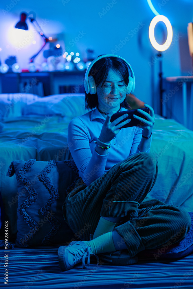 Teenage girl in wireless headphones sitting on floor playing video game in smartphone in dark bedroom Teenage girl in wireless headphones sitting on floor playing video game in smartphone in dark bedroom