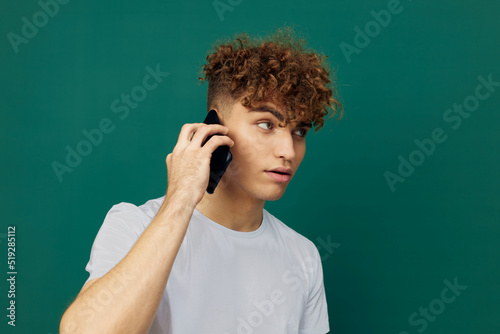 a funny, surprised man with curly hair stands against a green background and holds his black smartphone in his hand, talking on it, his mouth wide open in surprise © Tatiana