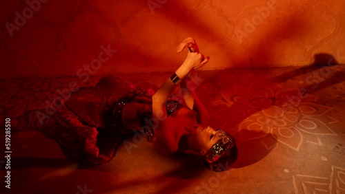 a woman with an adornment on head and in a sparkling bodice lies on the floor and moves hands beautifully against the wall with shadows. red light. the camera is moving photo