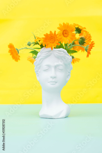 Creative concept colorful head of an ancient statue hair made of fresh flowers. Plaster sculpture of a female face on a yellow background. Vertical position