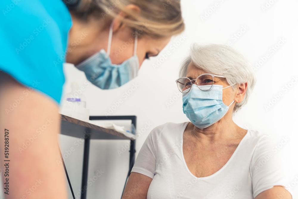 Fototapeta premium Senior lady with grey hair and her glasses on smiling sincerely at the doctor while wearing a face mask during a medical checkup. High-quality photo