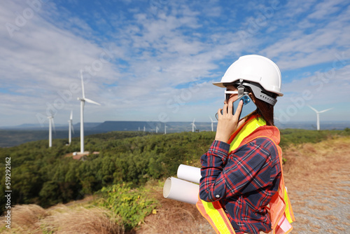 Windmill engineer young woman using mobile smartphone and planing renewable energy technology or alternative ecology project for future. Electrician engineer checking wind turbines site with blue sky