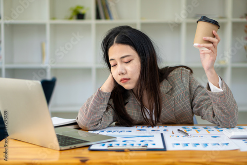 Sleepy Asian businesswoman yawning at work in the afternoon after being tired and fed up with working with laptop and financial graph documents at work.