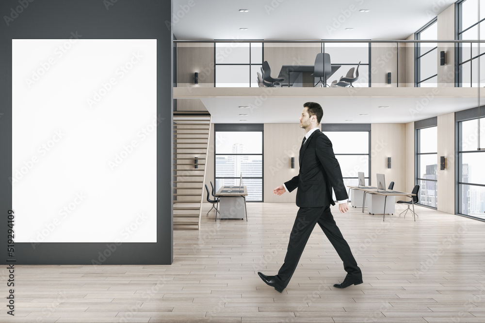 Side view of businessman walking in modern two floors wooden and concrete duplex office interior with empty white mock up poster on wall, panoramic city view, furniture and daylight.