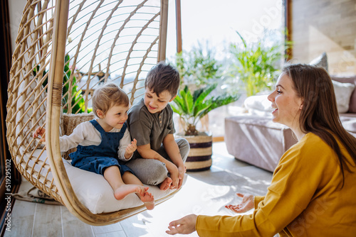 Young cheerful mother playing with her little children and having fun when swinging them on hanging chair in conservatory at home. photo