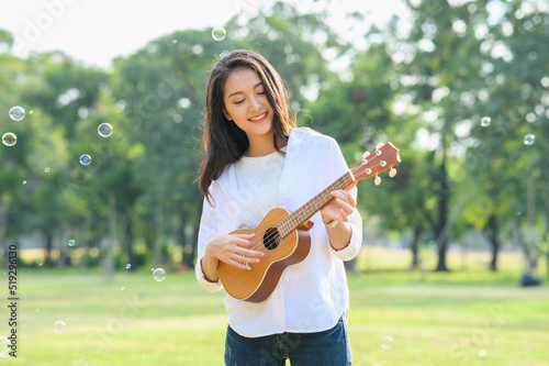 Young beautiful Asian woman playing ukulele in green park in the evening. Girl relaxing in outdoor park. photo