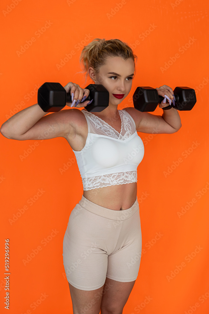 A beautiful young female trainer in a sports top holds pink dumbbells in her hands smiling.