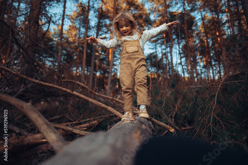 Autumn portrait of happy little girl balancing on tree trunk with arms outstretched outdoors in forest.
