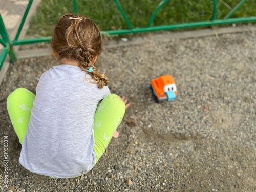 Little Caucasian girl with a cute ponytail and a blue hair binder in it plays on the gravel playground. A child having summer activity with their toy excavator on the sand photo