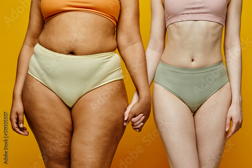 Close up of two real young women wearing underwear and holding hands against yellow background focus on different body shapes