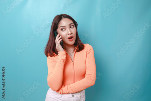A portrait of a surprised young Asian woman having a phone call, isolated on blue background