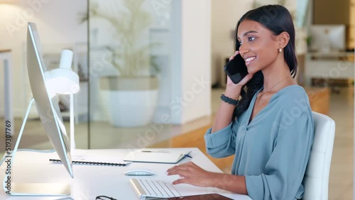 Woman answering a phone call in an office alone at work. One young and happy Indian corporate worker talking, having a conversation and typing a report or replying to emails on a desktop computer
