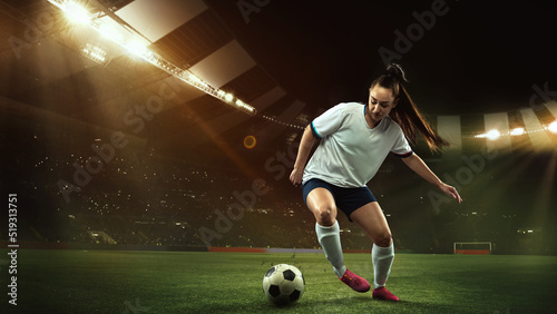 Female soccer, football player dribbling ball in motion at stadium during sport match over evening sky background.