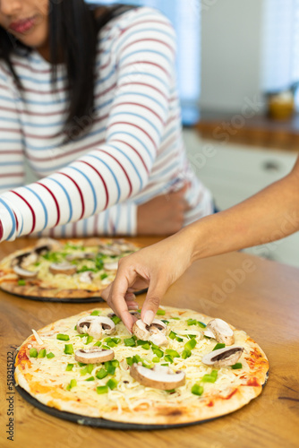 Hands of biracial female friends garnishing pizza with mushroom and bell peppers on kitchen island