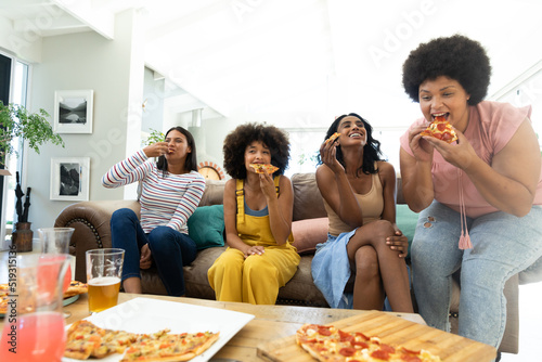 Happy female biracial friends enjoying pizza and drinks together in living room  copy space