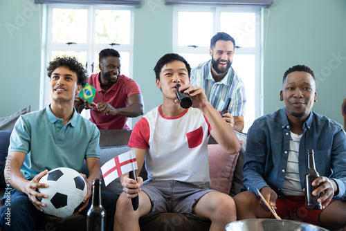 Happy multiracial male friends with ball and beer bottles watching soccer match on sofa at home