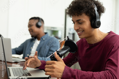 Smiling multiracial male podcaster speaking over microphone while recording podcast with coworker