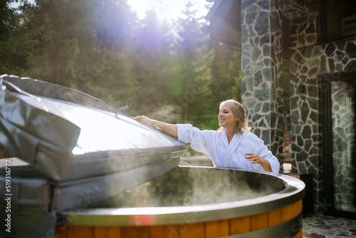 Fotografia Woman in bathrobe opening lid of hot tub, checking temperature, ready for home spa procedure in hot tub outdoors