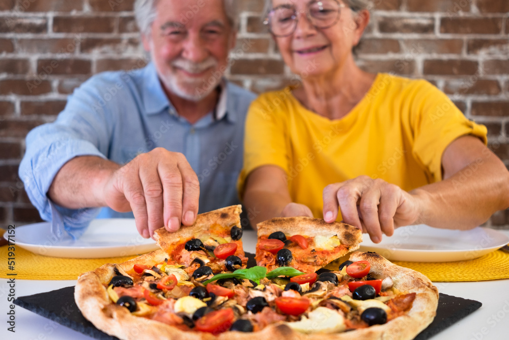 Defocused senior couple sitting at home table taking a slice of pizza enjoying delicious food Defocused senior couple sitting at home table taking a slice of pizza enjoying delicious food