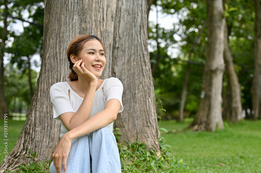 Attractive young Asian female relaxes sitting under a tree in the greenery of a park. Attractive young Asian female relaxes sitting under a tree in the greenery of a park.