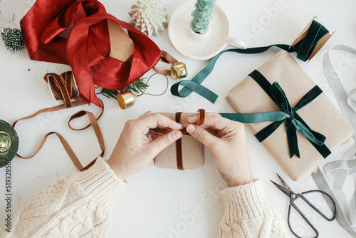 Hands wrapping stylish christmas gift flat lay. Person preparing simple christmas gift box, ribbons, craft paper and ornaments composition on white wood. Merry Christmas! Aesthetic winter holidays
