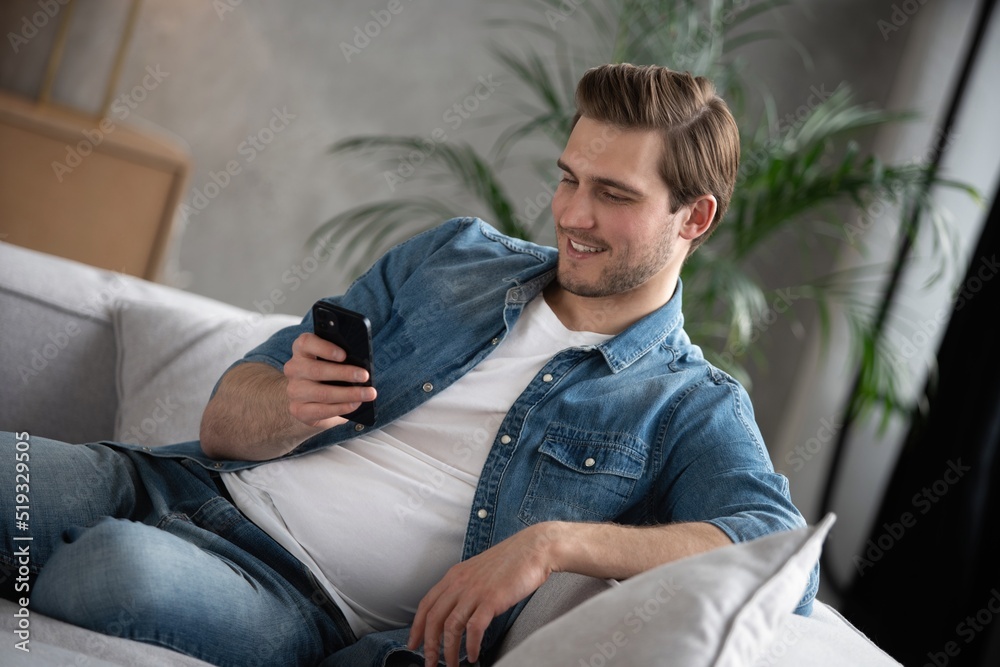 Fototapeta premium Attractive smiling young man wearing casual clothes sitting on a couch at the living room, using mobile phone