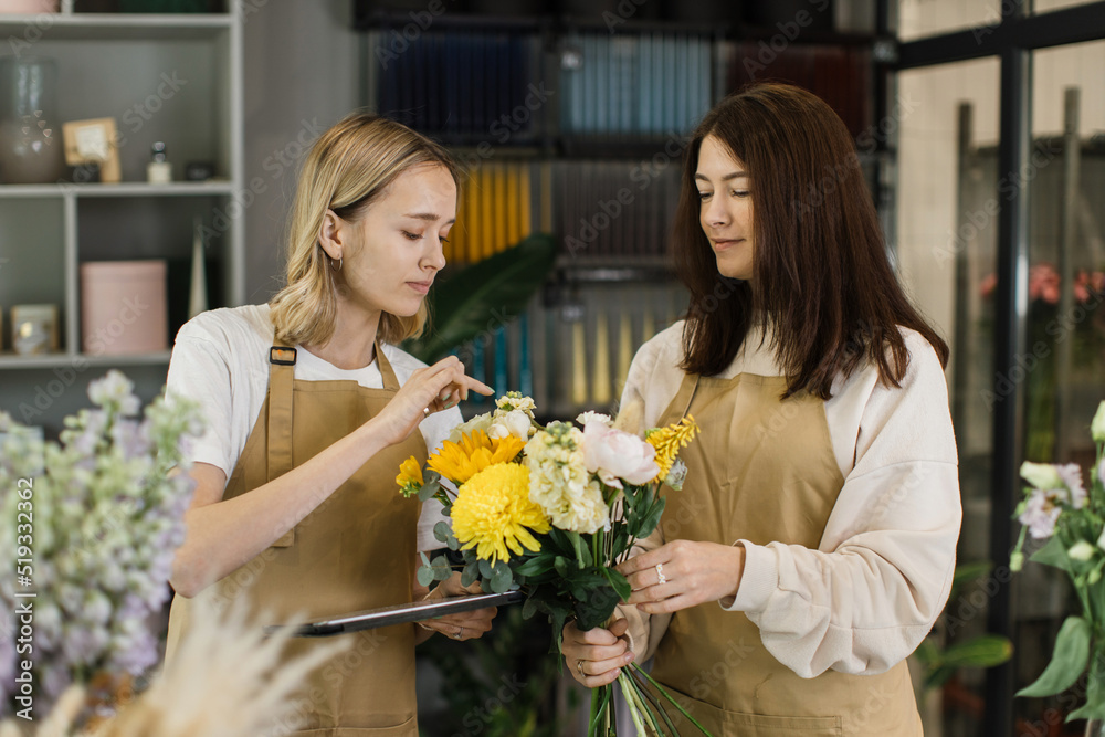 Team of two female florists in beige aprons using tablet while discussing which flowers to order for next week and making bouquet in modern interior floral shop. Team of two female florists in beige aprons using tablet while discussing which flowers to order for next week and making bouquet in modern interior floral shop.
