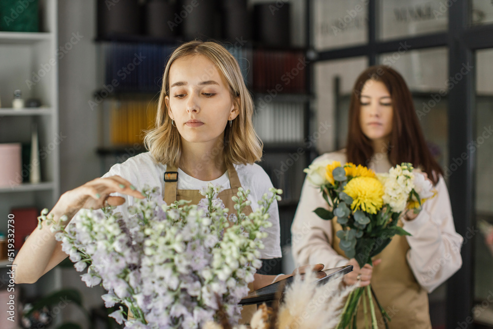 Two smiling female florists working behind counter in modern loft interior of floral shop. Floristics, business, decoration concept. Creative occupation in floral boutique.