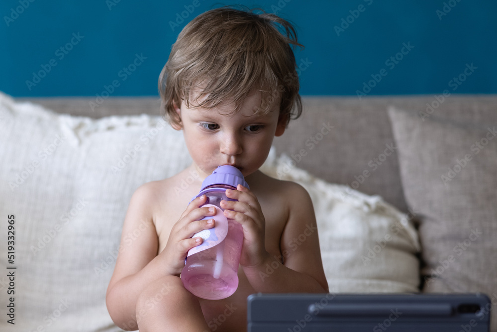 Cute blonde toddler child watches cartoons or playing games using a tablet pc, sitting on sofa at home and drinking water. Gadgets and modern entertainment for children. Video chat, video call. Cute blonde toddler child watches cartoons or playing games using a tablet pc, sitting on sofa at home and drinking water. Gadgets and modern entertainment for children. Video chat, video call.