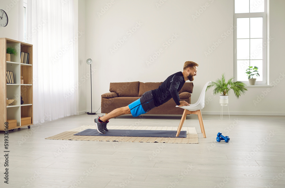 Gym at home. Athletic man is doing push up exercise with chair at home in his spacious living room with modern interior. Side view of motivated millennial guy training his muscles by doing sports.