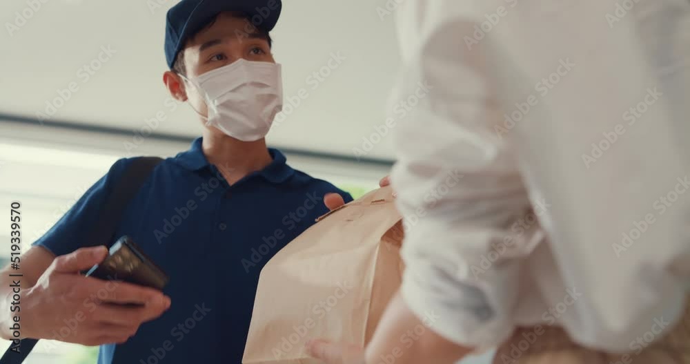 Close-up Young Asian delivery man wear mask in blue navy uniform carry case box knock door home online shopping to woman in front of door lady sign accept paper bag on smartphone at house concept. Close-up Young Asian delivery man wear mask in blue navy uniform carry case box knock door home online shopping to woman in front of door lady sign accept paper bag on smartphone at house concept.