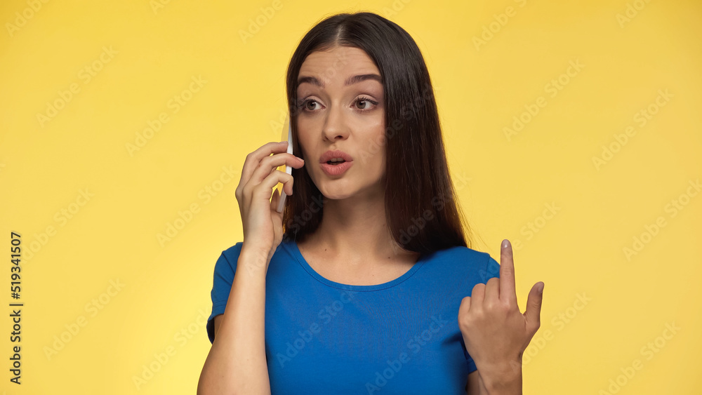 young brunette woman in blue t-shirt emotionally talking on smartphone isolated on yellow.