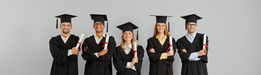 Happy proud diverse mixed race international college or university students in graduate hats and gowns holding paper diplomas and smiling at camera standing on gray background at graduation event
