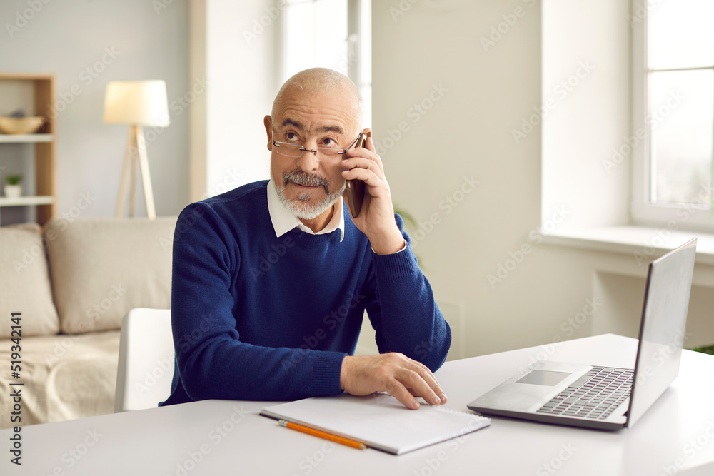 Serious mature businessman work on laptop at home office talk on cellphone call with business client. Focused middle-aged man boss or CEO use computer have smartphone communication online.