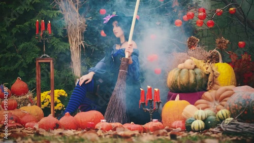 Medieval beautiful fantasy woman sits, holds mystical magic broom. Traditional pumpkin decor Halloween holiday. Blue vintage dress black cons hat, striped stockings. red candles burning, smoke flowing photo