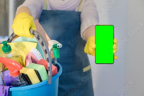 homeowner in rubber gloves holding detergent bottles. Hold mobile phone with blank empty green screen