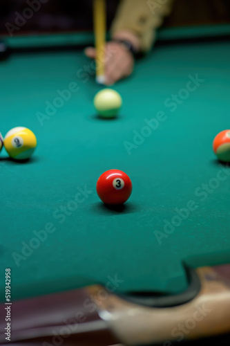 Close-up of a red ball number 3. A player's hand with a cue and balls on a green cloth on a blurred background.