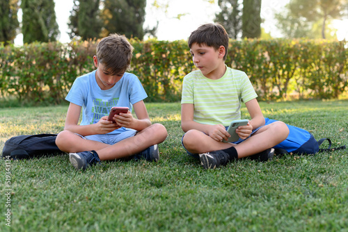 Two school children sitting in a park with their backpacks and mobile phones.