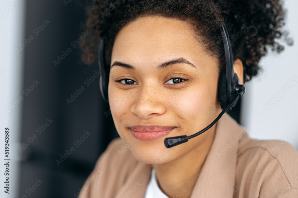 Close-up photo of pretty, confident curly-haired young african american woman with headset, call center worker or consultant, wearing stylish shirt, looks directly at the camera, smiling Close-up photo of pretty, confident curly-haired young african american woman with headset, call center worker or consultant, wearing stylish shirt, looks directly at the camera, smiling