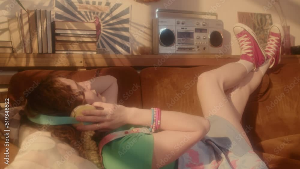 Full side view of red-haired Caucasian girl in over-ear headphones listening to music via tape player, lying on couch in living room at daytime, having fun