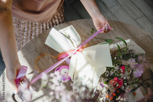 a woman florist ties a pink ribbon bouquet wrapped in paper. The bouquet lies on a round wooden table. Top view