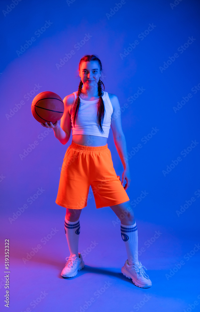 a girl with a basketball in orange shorts, a t-shirt and a baseball cap. sportswoman in golfs and sneakers on a blue background. Physical education teacher. basketball player a girl with a basketball in orange shorts, a t-shirt and a baseball cap. sportswoman in golfs and sneakers on a blue background. Physical education teacher. basketball player