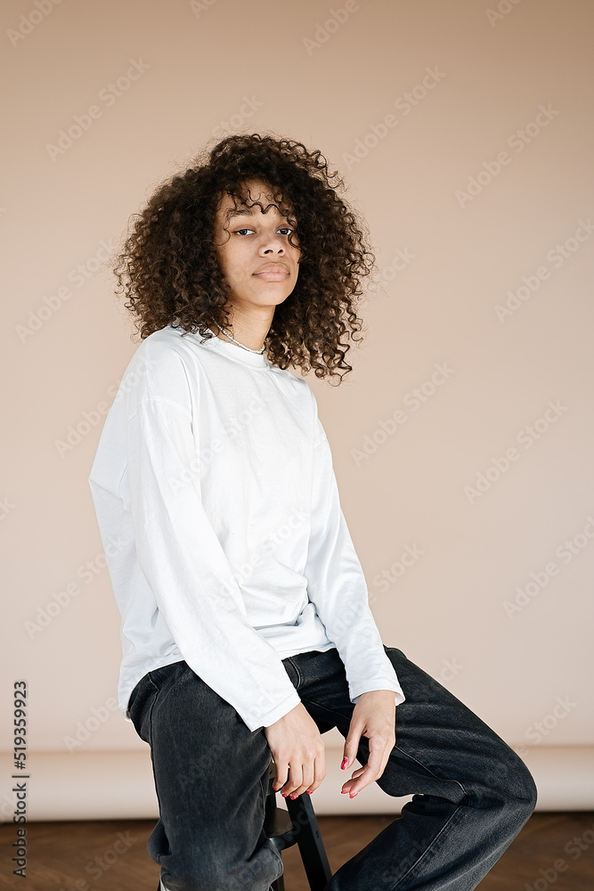 Studio portrait african american girl sitting on a chair in a wearing casual white t-shirt isolated beige background 