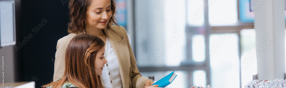 Fototapeta premium smiling woman showing new copybook to daughter in stationery store, banner.