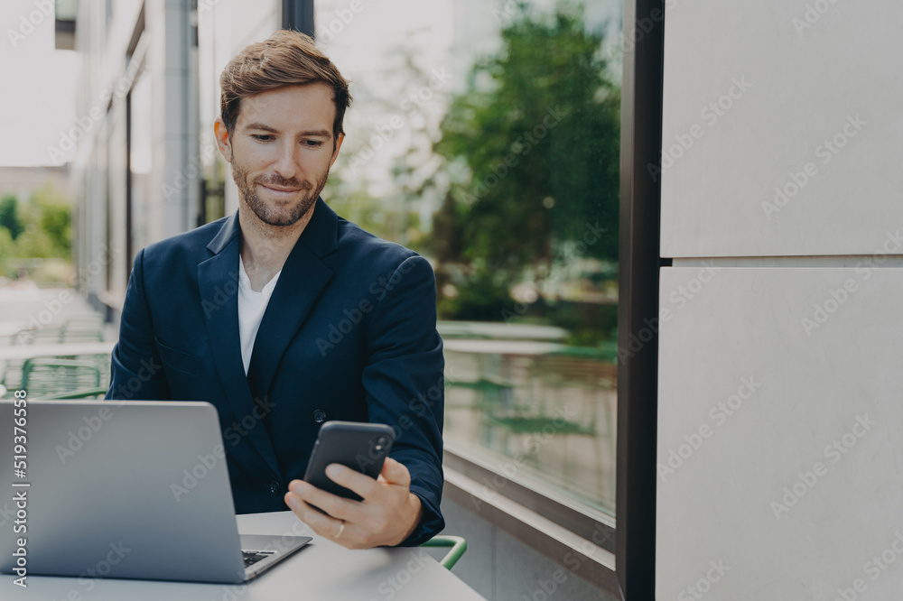Focused young businessman working on laptop in outdoor cafe and browsing phone notifications Focused young businessman working on laptop in outdoor cafe and browsing phone notifications