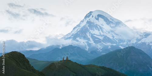 Small silhouette of Gergeti Trinity Church XIV century with a huge Kazbek 5054m mountain. Landscape shot from the Stepantsminda village in Georgia.